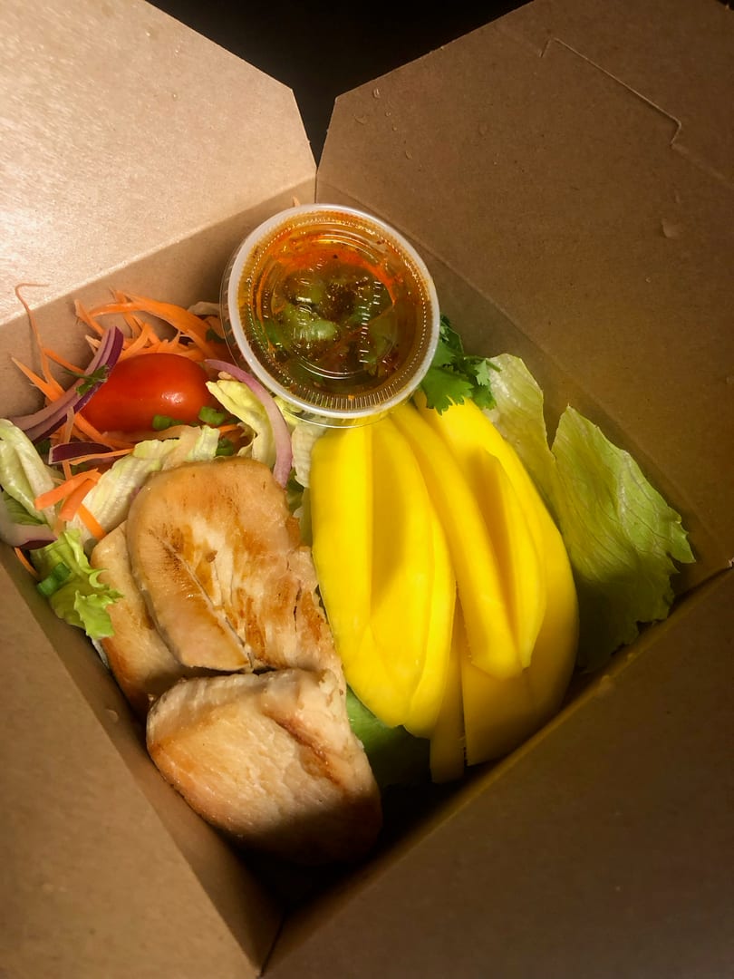 Grilled chicken, mango slices, and salad with dressing in a brown takeout box.
