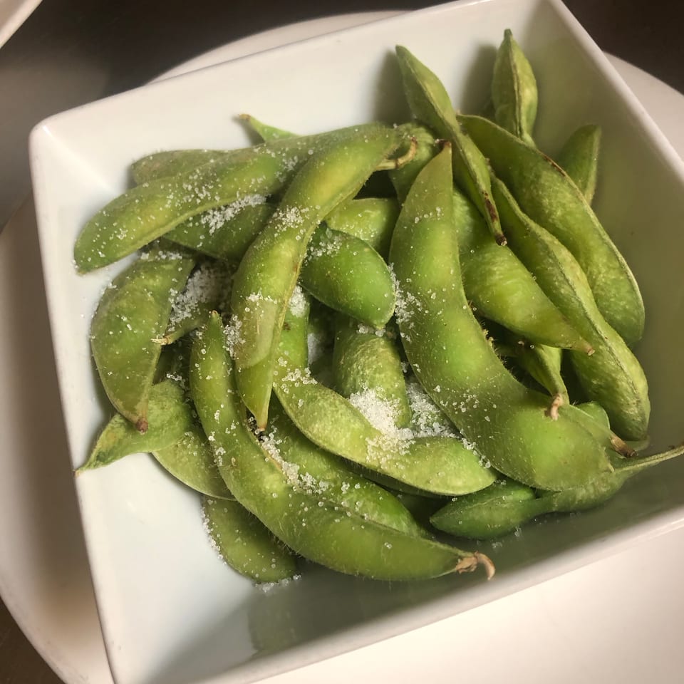 Steamed edamame pods sprinkled with coarse salt in a white square bowl on a restaurant table.